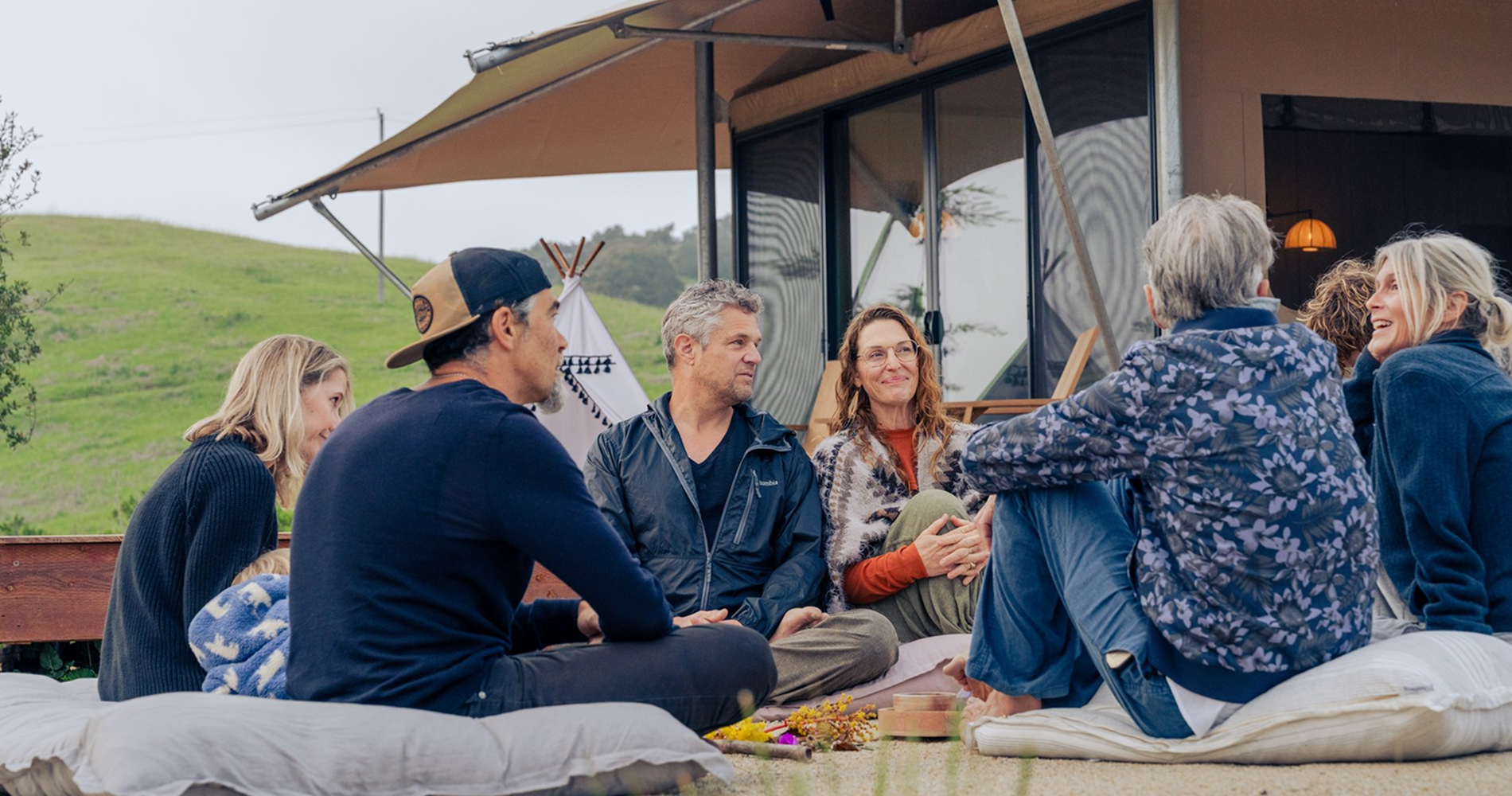 A group of eight people sits casually on cushions outside a modern building, engaged in conversation with a green hillside in the background.