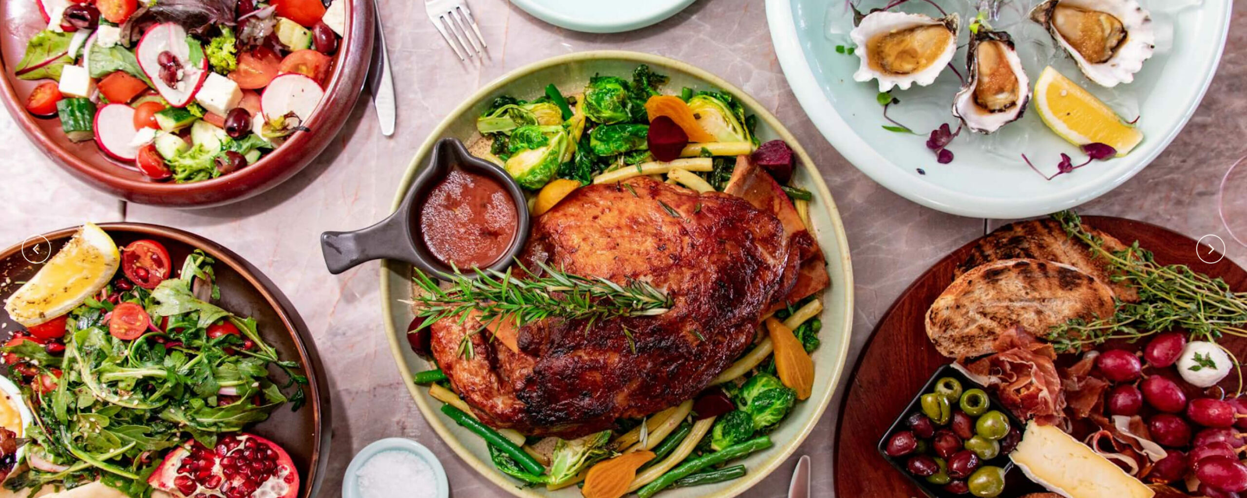A variety of dishes including a roasted meat with vegetables, salads, fresh oysters, and a cheese platter.