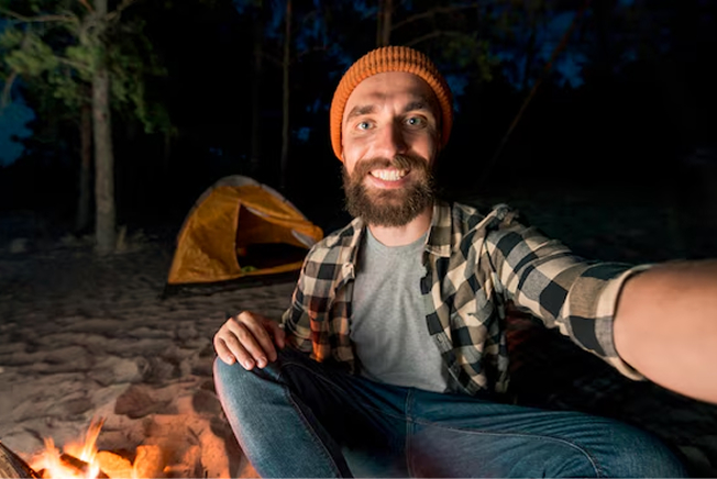 A man with a beard and orange beanie smiles for a selfie by a campfire, with a yellow tent visible in the background.