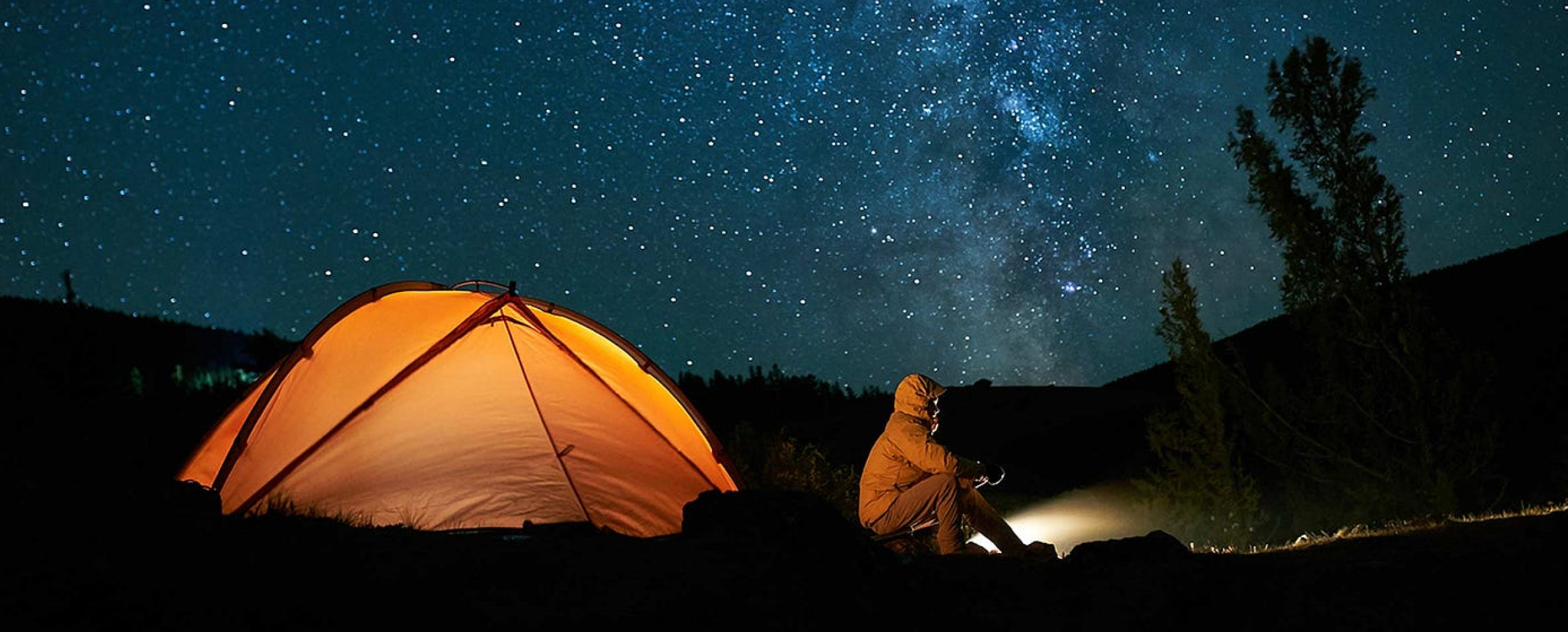 An orange tent is set up under a starry night sky, with a person sitting nearby, holding a flashlight.