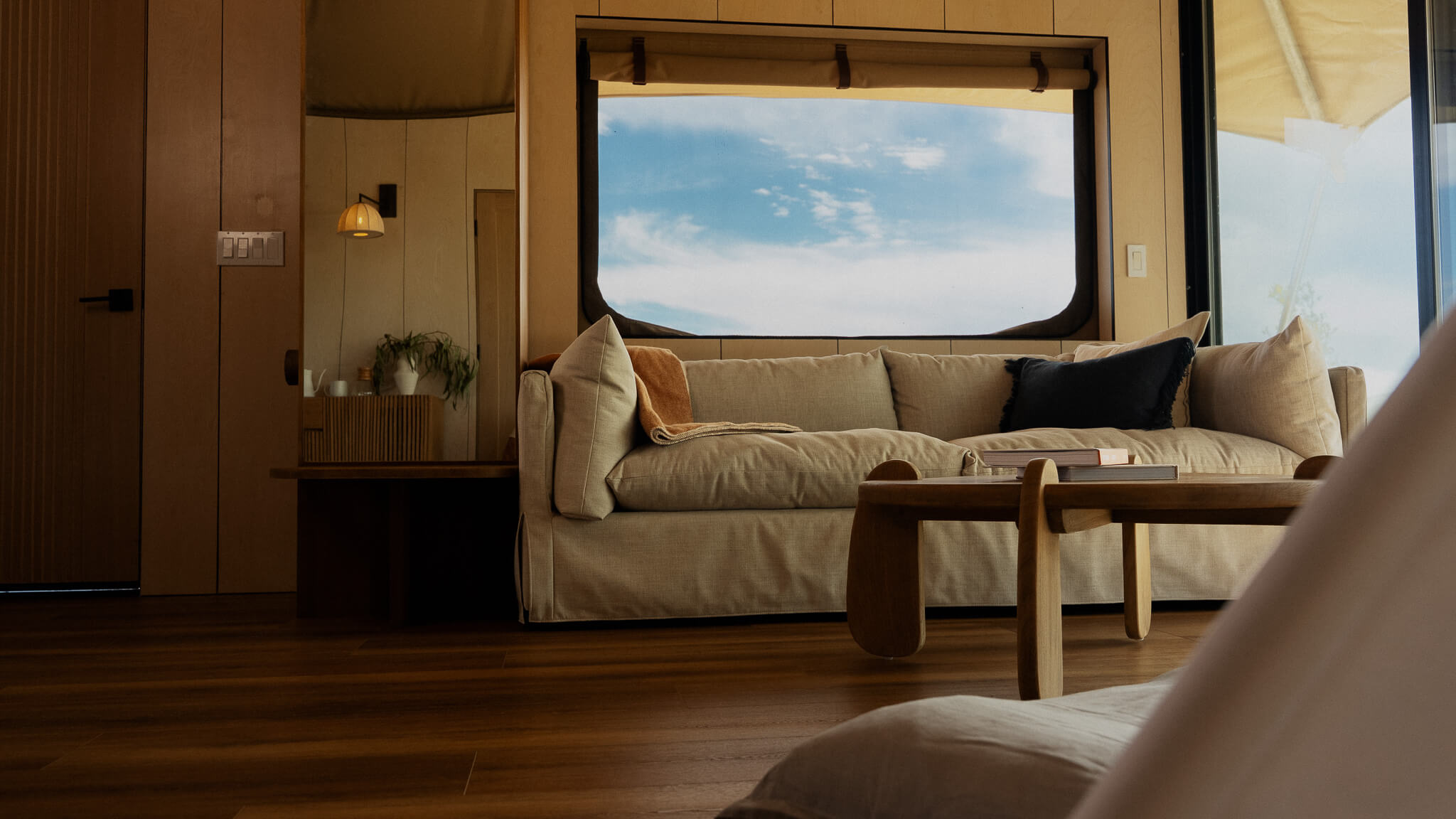 A modern, cozy living room features a light-colored sofa with pillows, a wooden coffee table, and large windows exposing a blue sky.