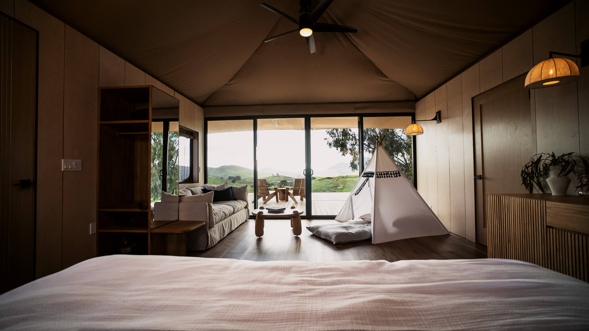 A cozy interior of a tent-style room featuring a bed, seating area, and large windows with scenic views.