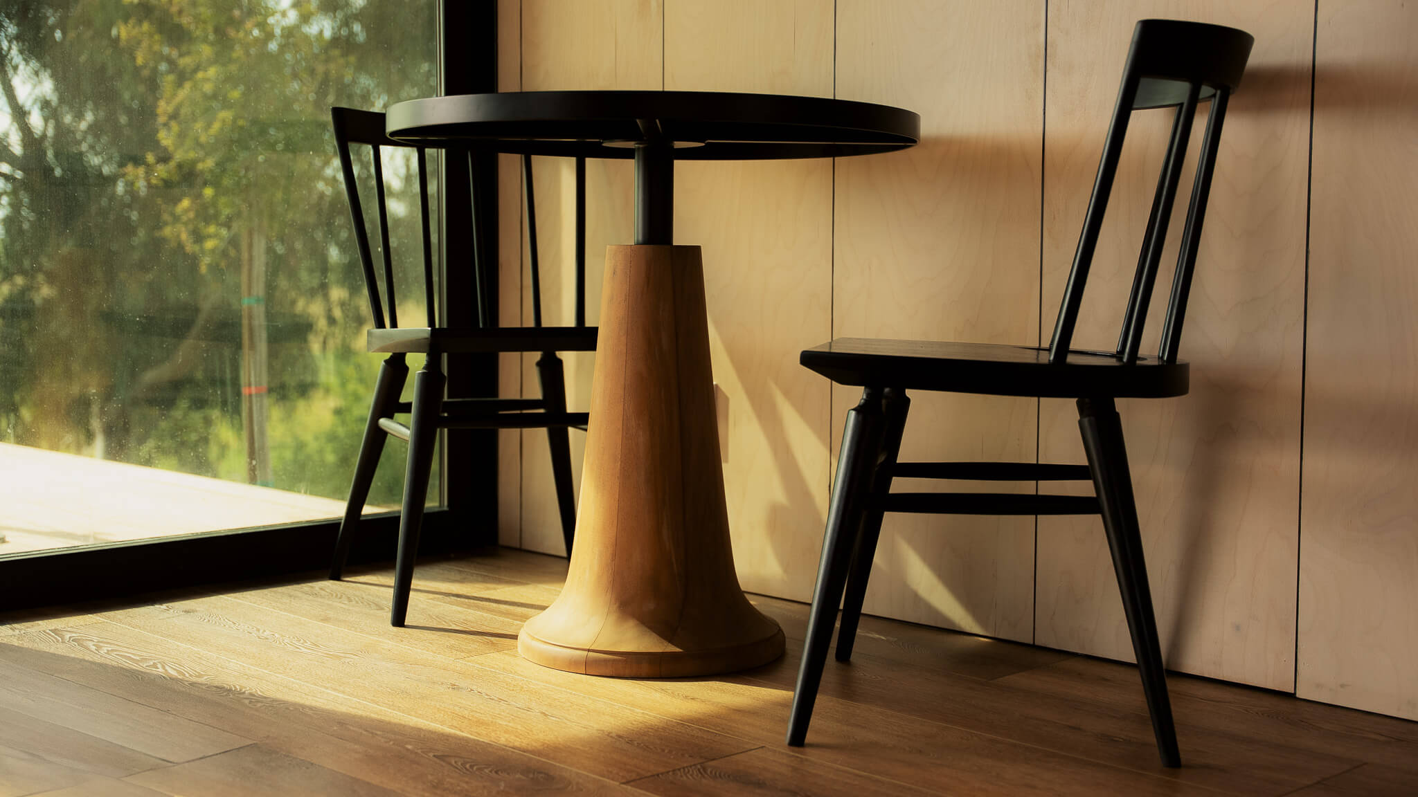 A small black round table with a unique wooden base, flanked by two black chairs, in a well-lit interior setting.