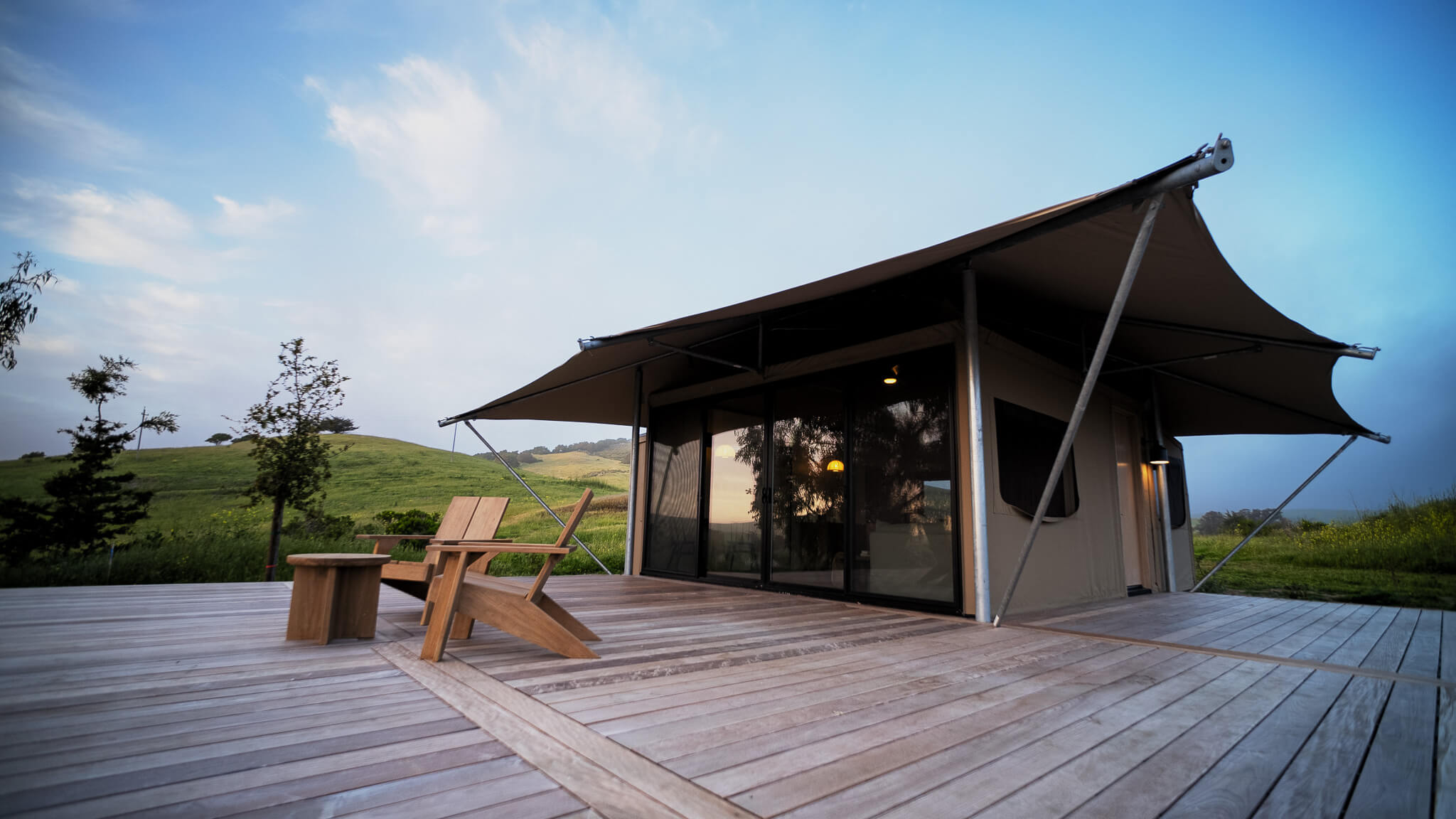 Modern cabin with a unique roof design on a wooden deck, surrounded by green hills under a partly cloudy sky.