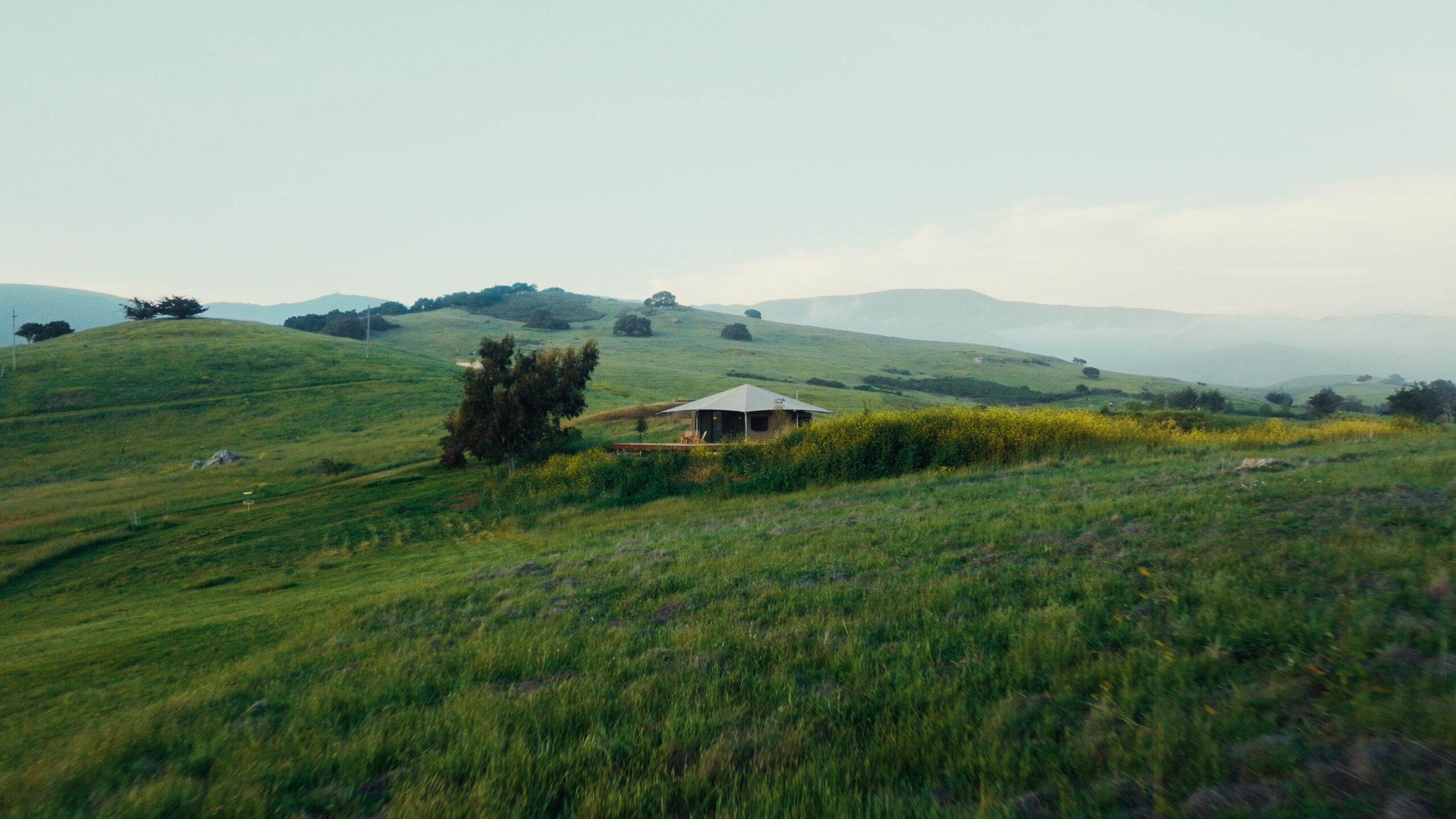 Lush green hills stretch into the distance, featuring a solitary house surrounded by vegetation under a light blue sky.