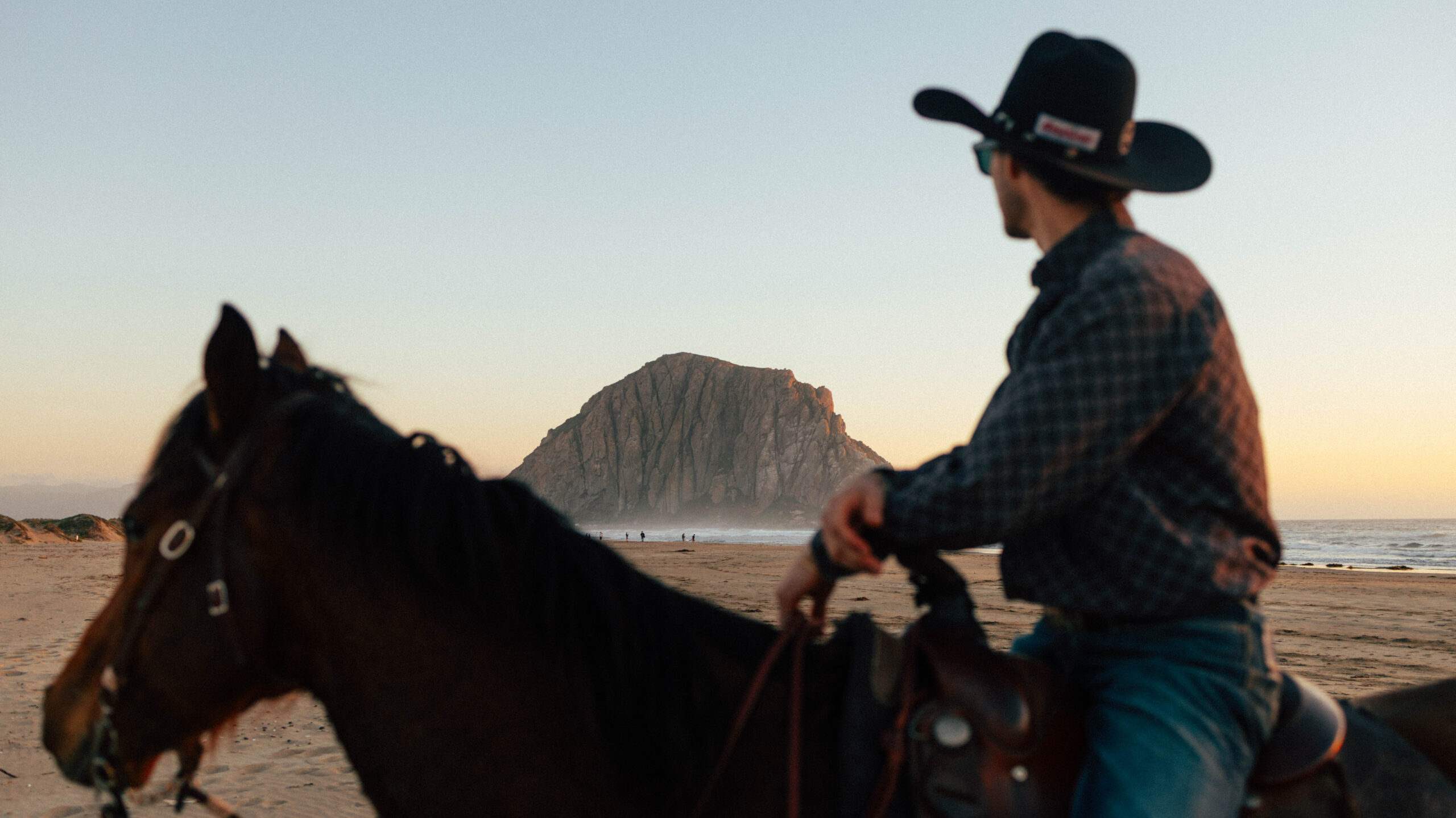 A person in a cowboy hat sits on a horse at the beach, facing a large rock formation at sunset.