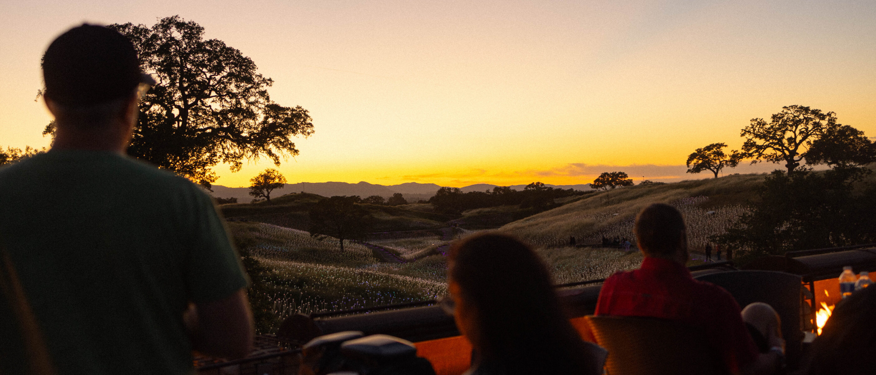 People enjoy a sunset view over rolling hills, with silhouettes against a colorful sky transitioning from orange to purple.