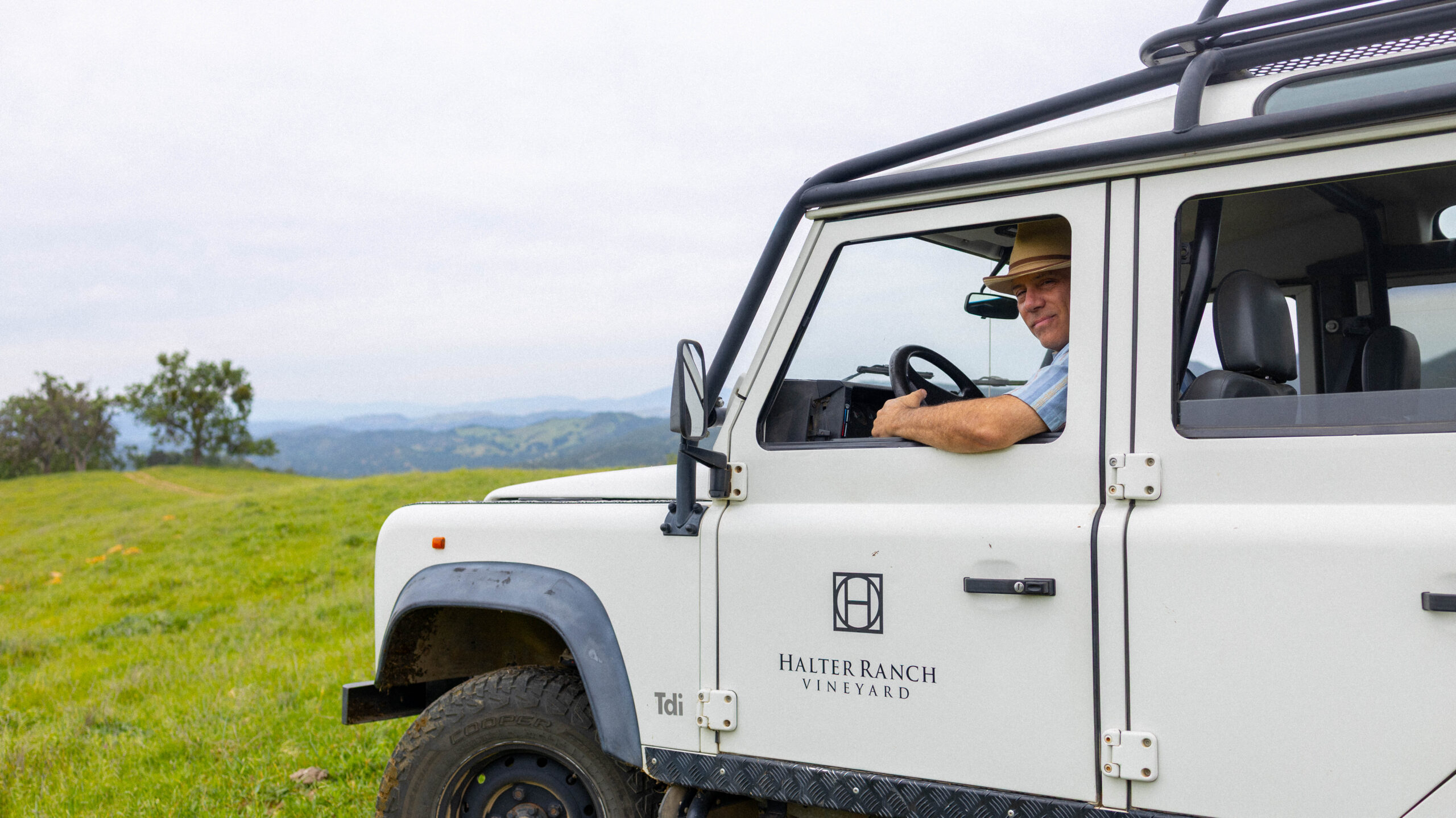 A person wearing a hat sits in the driver's seat of a white vehicle on a grassy hillside with mountains in the background.
