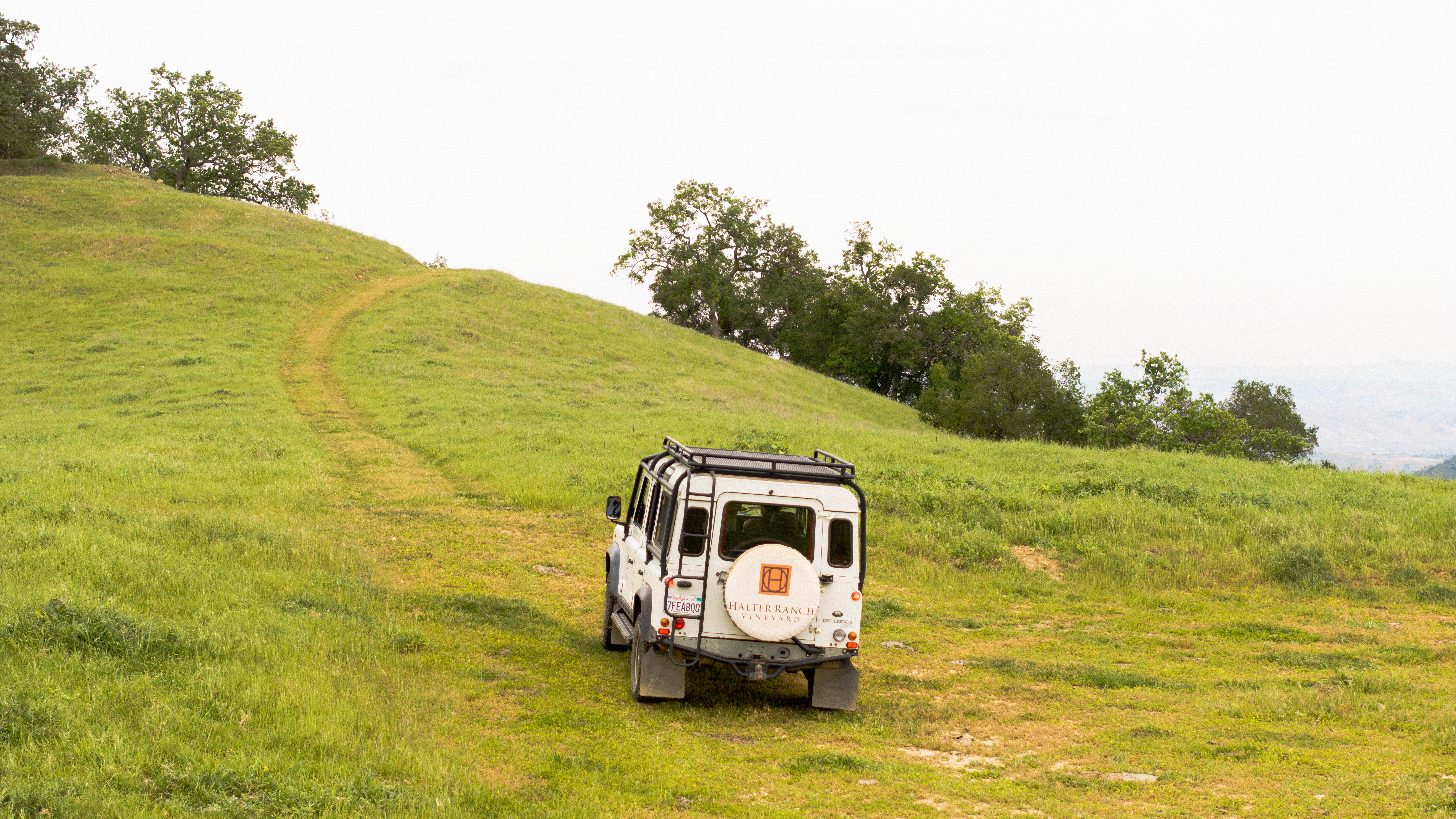 A white off-road vehicle drives along a dirt path through a green, hilly landscape under a cloudy sky.