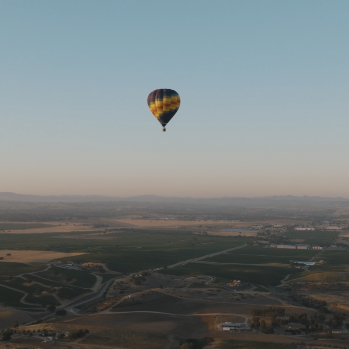 A colorful hot air balloon floats above a wide landscape of fields and hills under a clear sky.