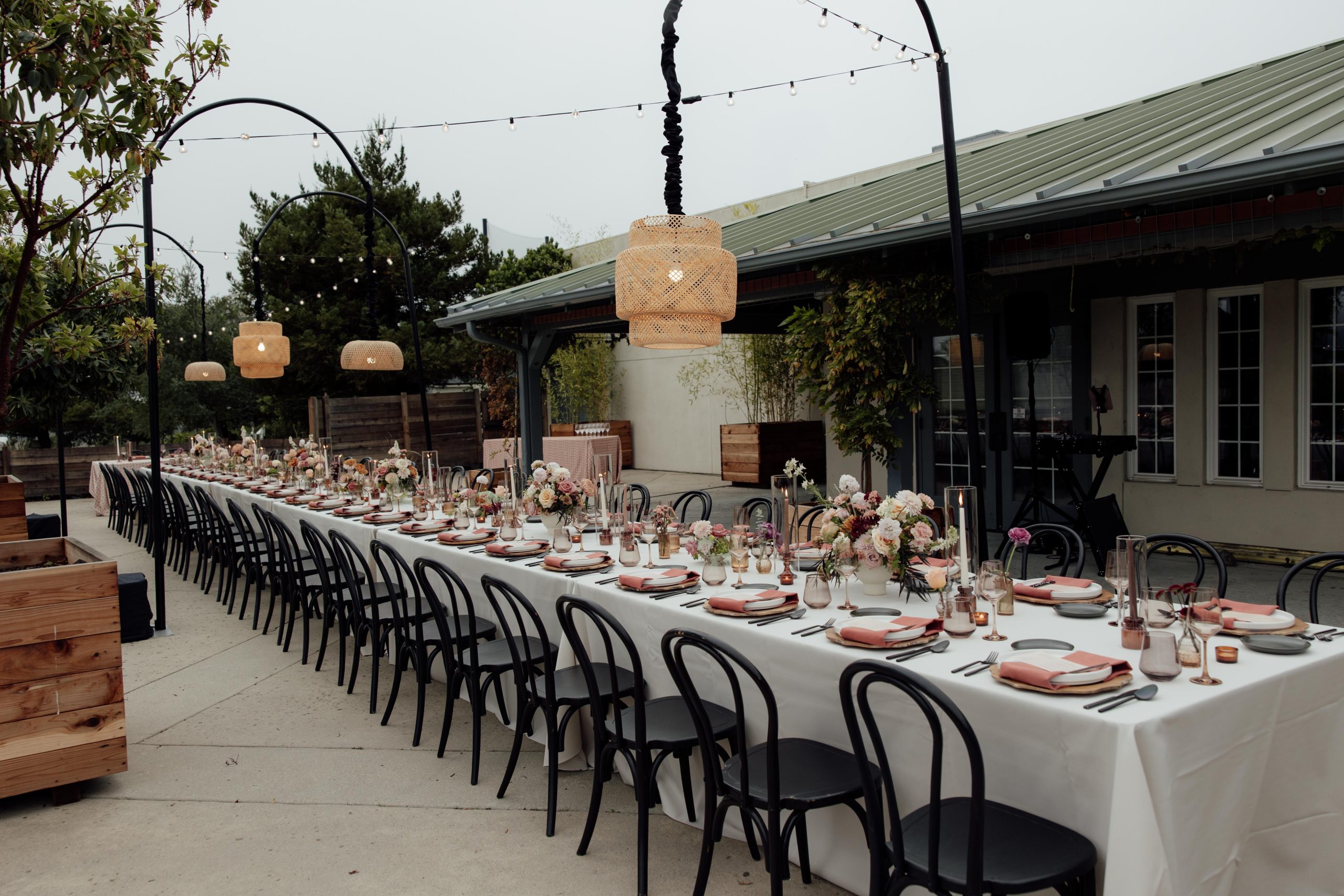 A long outdoor dining table is set for a gathering, adorned with flowers and tableware, under hanging lights.