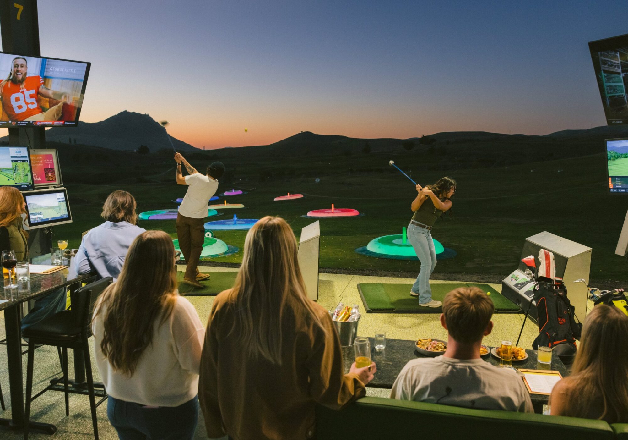 A group of people observe a golfer taking a swing at a driving range with colorful targets, set against a sunset backdrop.