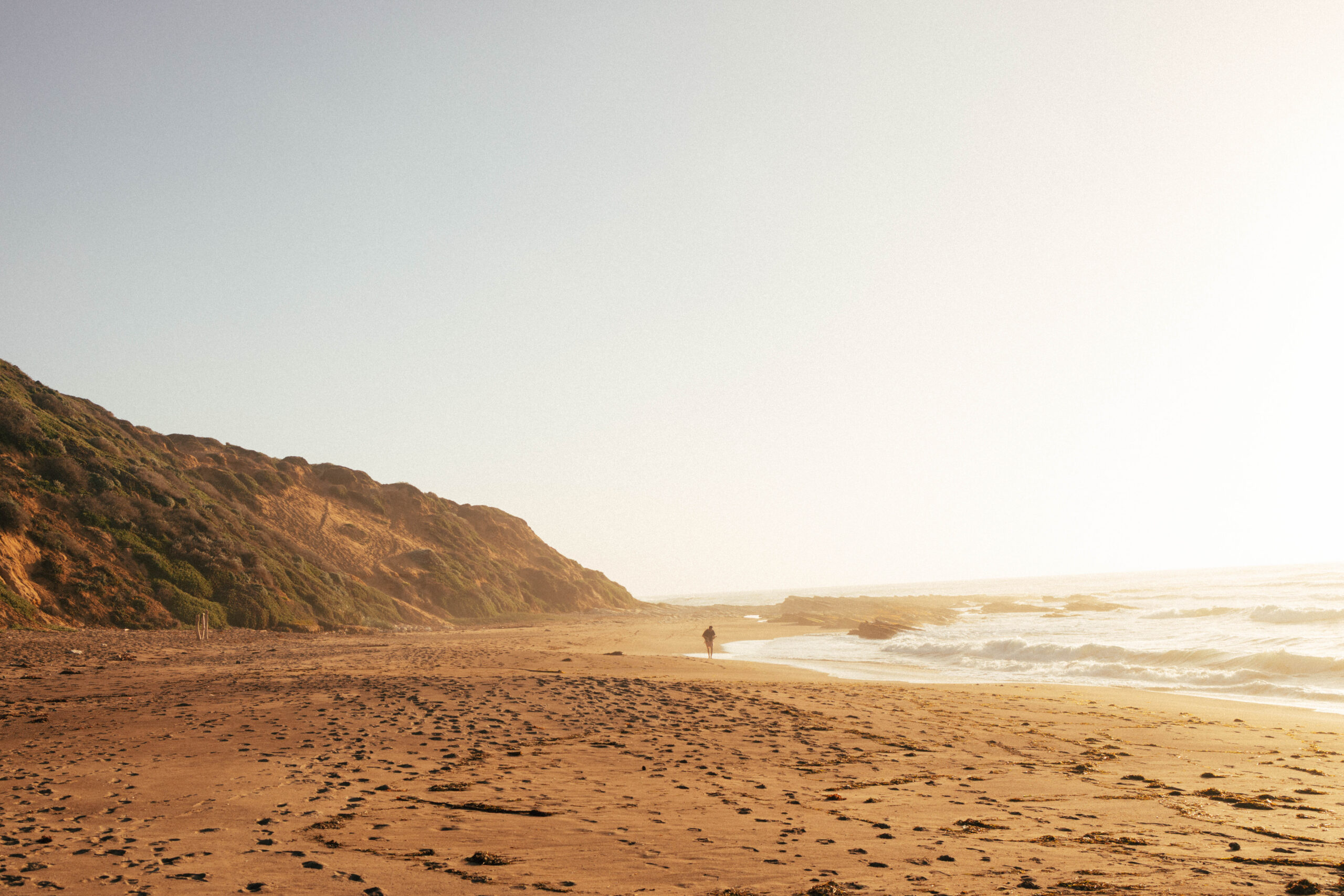 A person walks along a sandy beach with gentle waves, framed by cliffs and a hazy sky during sunset.