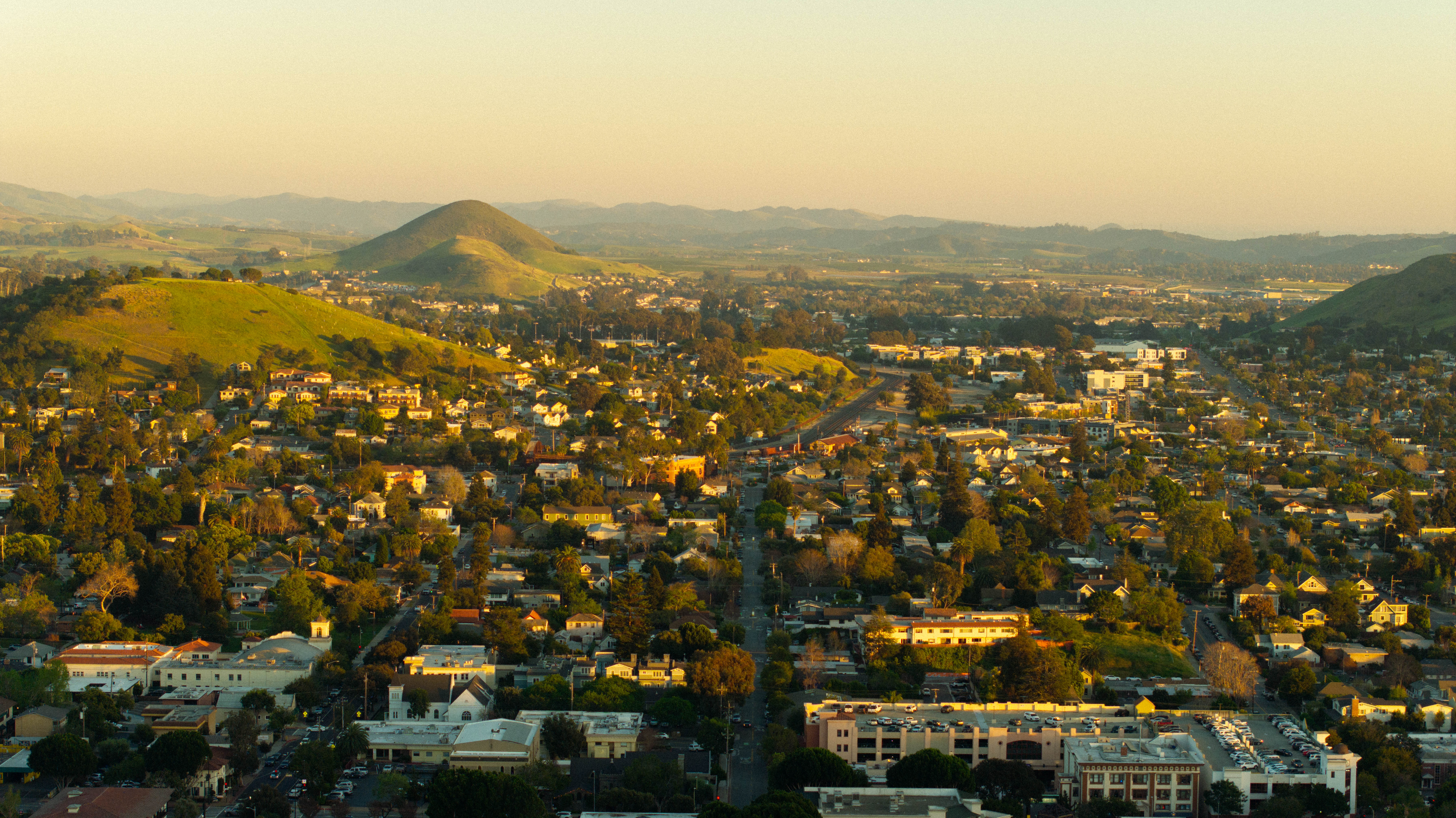A panoramic view of a town surrounded by rolling hills during sunset, showcasing residential areas and distant mountains.