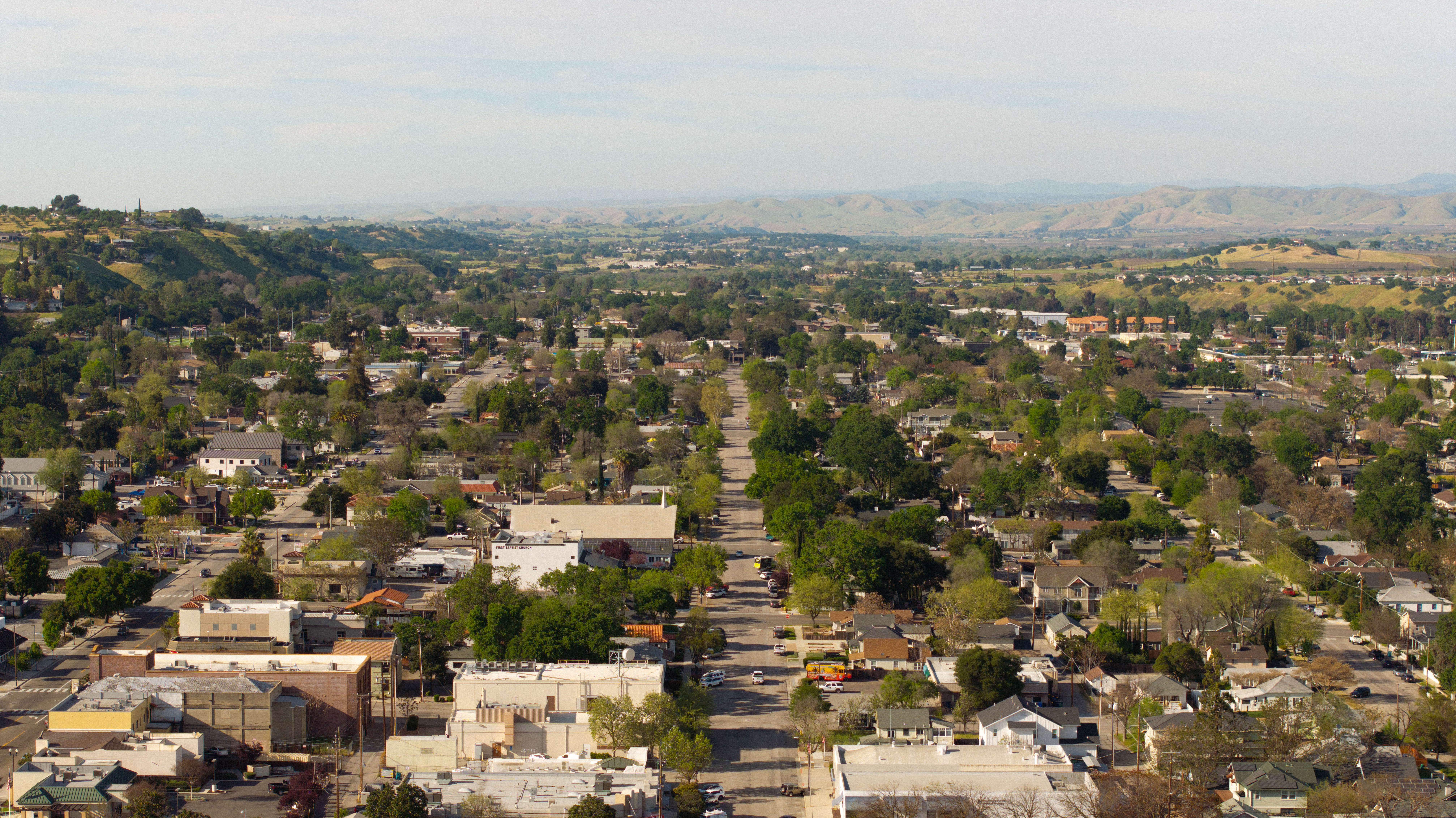 Aerial view of a suburban area with tree-lined streets, houses, and distant mountains under a clear sky.