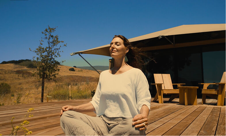 A woman practices meditation outdoors on a wooden deck, with blue skies and hills in the background.